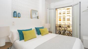 Une chambre d’hôtel avec un lit, une table de chevet, une lampe, un miroir et un balcon donnant sur des immeubles.