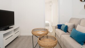 Ett modernt vardagsrum med en platt-TV, två sidobord i rotting, en beige soffa med blå kuddar och en vit vägg med en spegel.