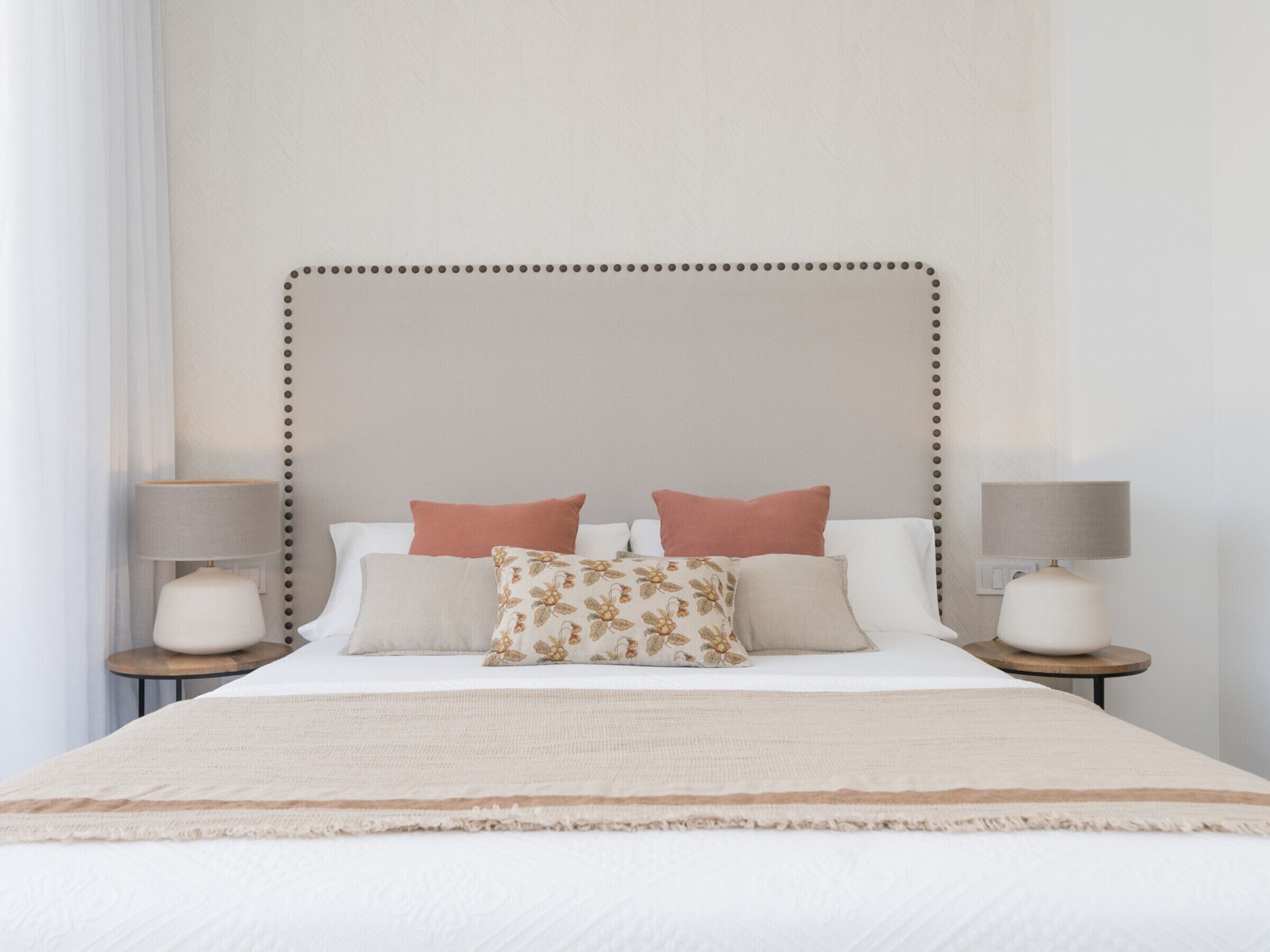 A neatly made bed with a beige headboard, two bedside tables with lamps, and a window with white curtains.