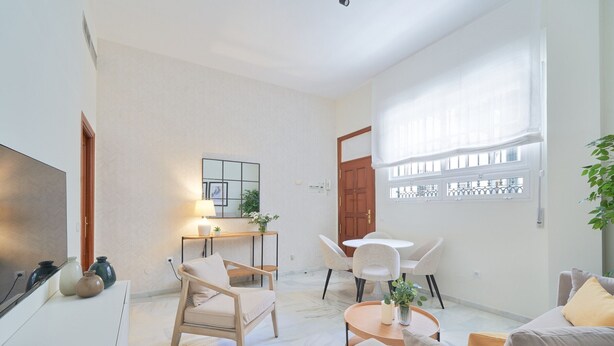 A modern living room with a sofa, chairs, a coffee table, and a TV. There is a desk with a lamp and a mirror. The room has a large window with curtains. - Elegant 2-Bedroom Apartment in Central Málaga, Close to Key Attractions (Málaga)