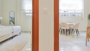 A modern bedroom with a bed, bedside table, and a mirror. There is a dining area with a table and chairs, and a window with blinds. - Elegant 2-Bedroom Apartment in Central Málaga, Close to Key Attractions (Málaga)