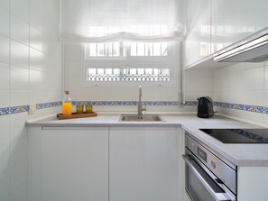 Eine Küche mit weißen Fliesenwänden, einem Fenster mit Schmiedeeisen-Gitter, einem Spülbecken, einer Kaffeemaschine und einem Backofen.