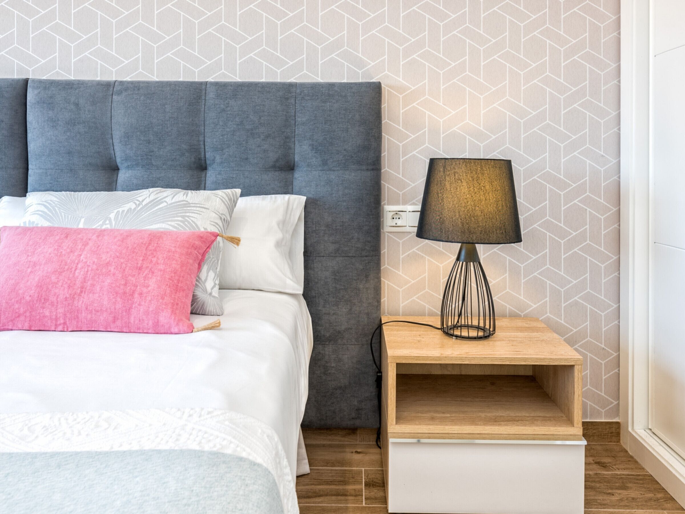 Un lit bien fait, avec une tête de lit grise, des oreillers blancs et roses, et une table de chevet en bois sur laquelle se trouve une lampe.