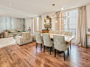 Ein geräumiges Wohnzimmer mit Holzboden, einem beigefarbenen Sofa, einem Glastisch und einem Essbereich mit weißem Tisch und grau gepolsterten Stühlen.