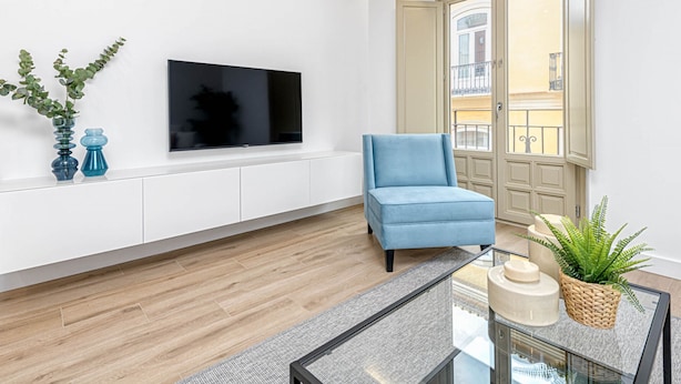 A modern living room with a flat-screen TV mounted on the wall, a blue armchair, a glass coffee table, and a view of the outside balcony. - Charming Apartment Near Alcazaba Málaga with Modern Amenities, WiFi, and A/C (Málaga)