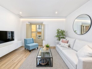 Ein modernes Wohnzimmer mit Flachbildfernseher, blauem Sessel, Glastisch, grauem Sofa und einem großen Spiegel.