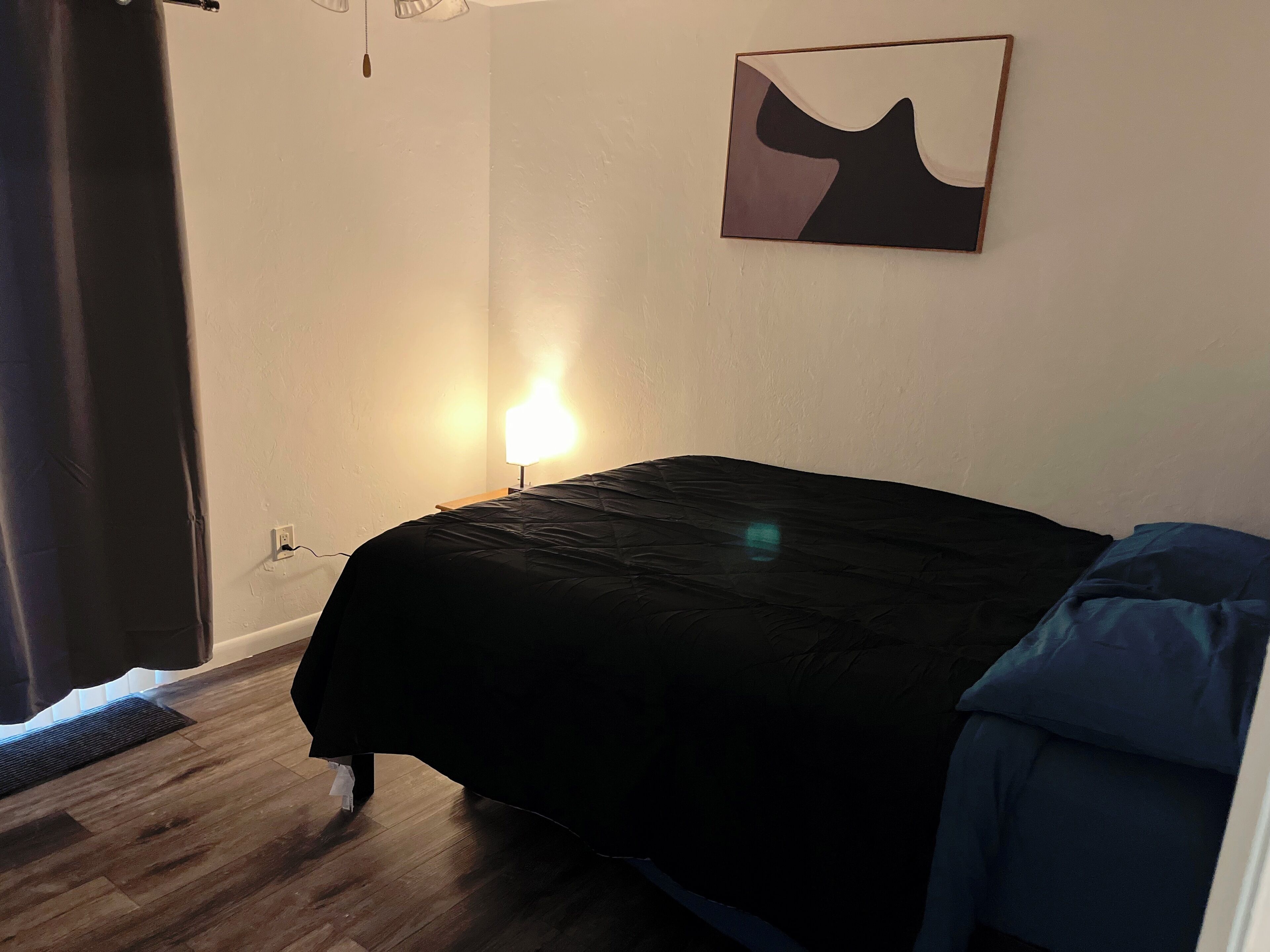 Un dormitorio con cama, mesita de noche, lámpara, un cuadro y ventana con cortinas.