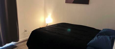 Un dormitorio con cama, mesita de noche, lámpara, un cuadro y ventana con cortinas.