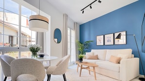 Una sala de estar moderna con un sofá blanco, sillones color beige, una mesa de comedor redonda y un ventanal con vistas a edificios.