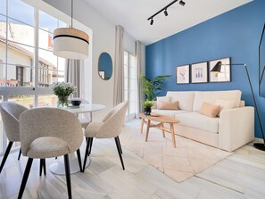 Una sala de estar moderna con un sofá blanco, sillones color beige, una mesa de comedor redonda y un ventanal con vistas a edificios.