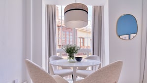 Una habitación moderna con una mesa redonda blanca, dos sillas color beige, una lámpara colgante, un jarrón con flores y vistas a edificios a través de grandes ventanales.