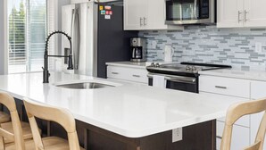 Une cuisine moderne avec des meubles blancs, un grand îlot central doté de tabourets, des appareils en acier inoxydable et un pare-feu en carreaux.