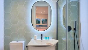 Ein Badezimmer mit sechseckigen Fliesen an der Rückwand, einem runden Spiegel, einer Toilette, einem Waschbecken mit Pflegeprodukten und einer Duschkabine aus Glas.