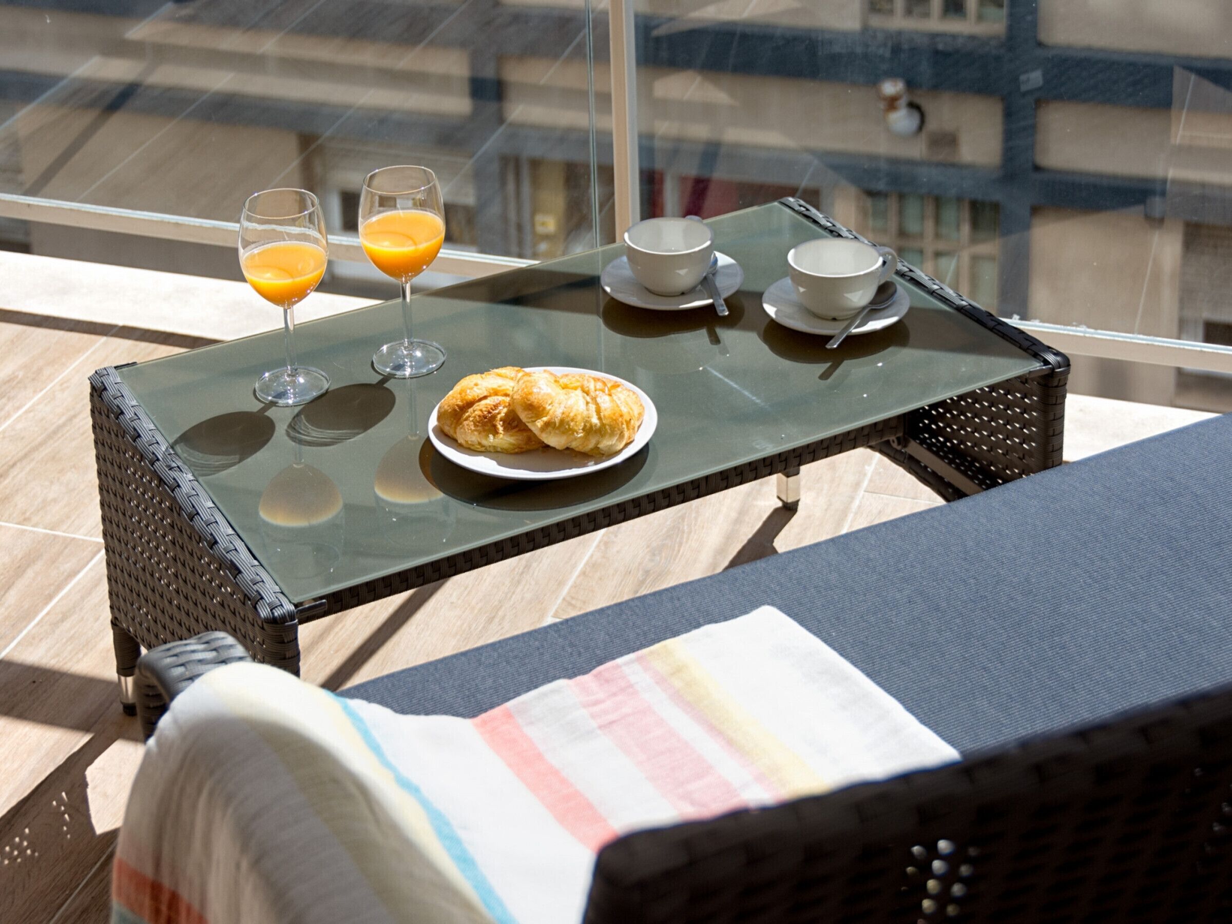 A balcony with a glass table set with croissants, two glasses of orange juice, and a cup and saucer.