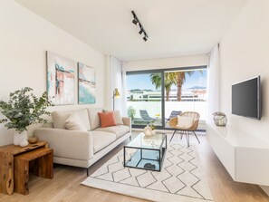 Een moderne woonkamer met een bank, salontafel en stoelen, een groot raam met uitzicht op palmbomen en gebouwen, en een flatscreen televisie aan de muur.