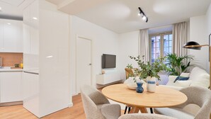 A modern living room with a round wooden table, four grey upholstered chairs, a white sofa, a TV on the wall, and a large window with curtains. - Charming 2-Bedroom Apartment in Central Malaga with Modern Amenities (Málaga)