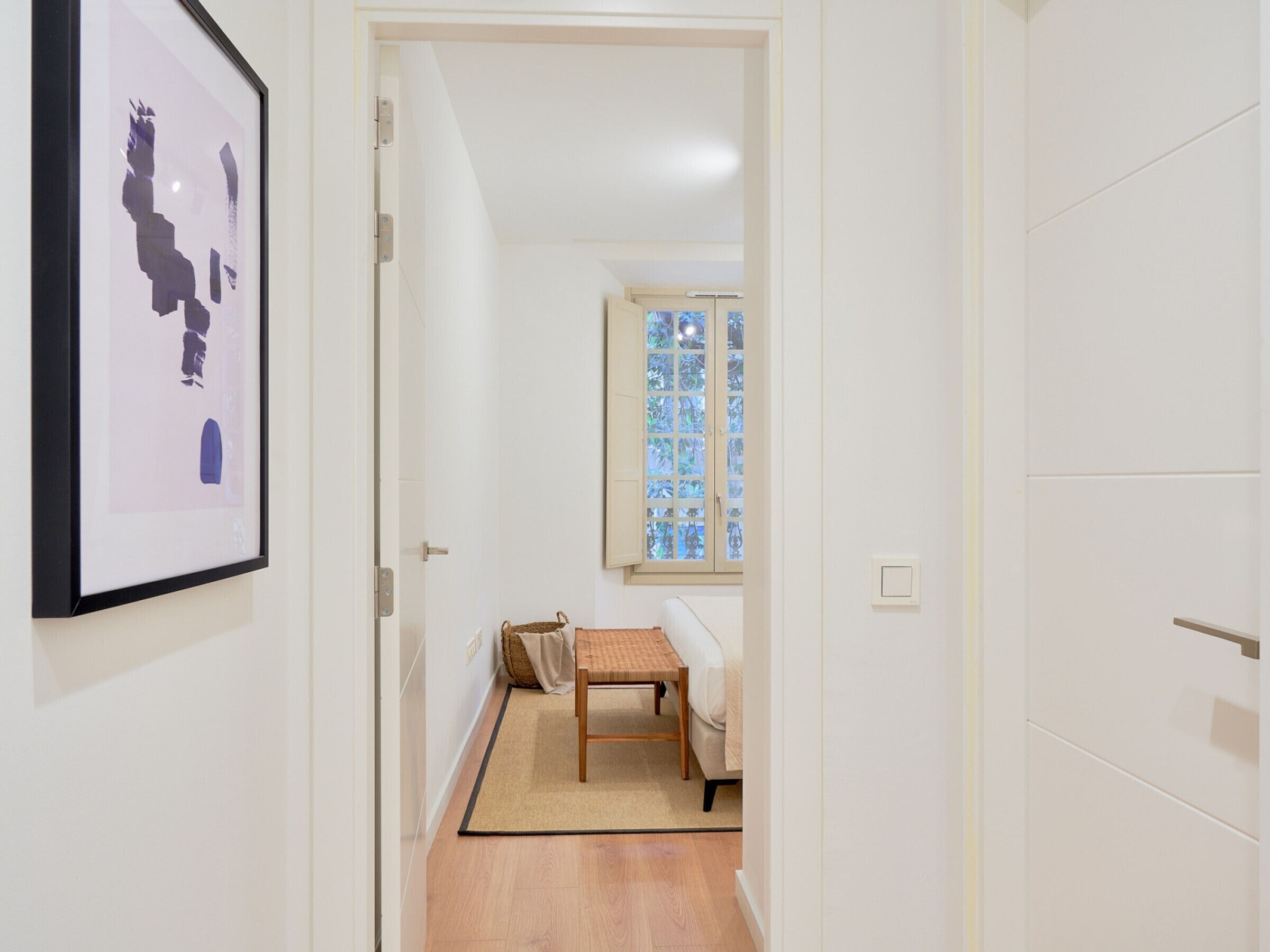 A hallway with a framed abstract artwork, a wooden bench, and a window with a stained glass design. - Charming 2-Bedroom Apartment in Central Malaga with Modern Amenities (Málaga)