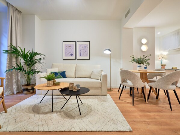 Un moderno salón con un sofá, una mesa de centro y sillas. Hay dos cuadros en la pared, una lámpara de pie y una planta en maceta.