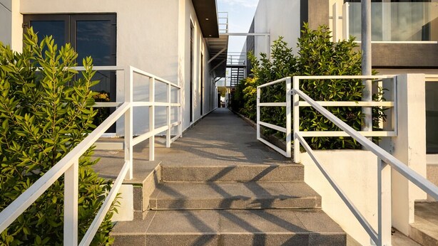Un bâtiment moderne avec un escalier menant à une porte, entouré de verdure et de rampes blanches.