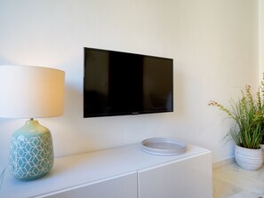 Ein modernes Wohnzimmer mit einem an der Wand montierten Flachbildfernseher, einer dekorativen Lampe auf einem weißen Schrank und einem Blumentopf auf dem Boden.
