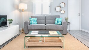 Een moderne woonkamer met een grijze bank, een glazen salontafel en een televisie op een witte kast.