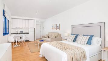 Een moderne, goed verlichte slaapkamer met een groot bed, nachtkastjes en uitzicht op een keuken en woonruimte.