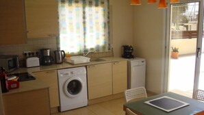 A compact kitchen with a washing machine, a toaster, a coffee maker, and a small refrigerator. There is a dining table with chairs and a window with curtains. - Athena Villa (Ayia Napa)