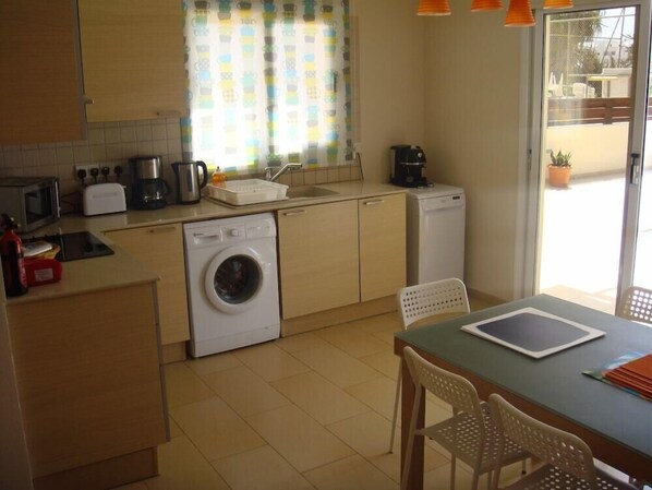 A compact kitchen with a washing machine, a toaster, a coffee maker, and a small refrigerator. There is a dining table with chairs and a window with curtains. - Athena Villa (Ayia Napa)