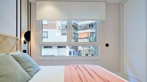 Een slaapkamer met een bed, een raam met uitzicht op gebouwen, en een wandlamp.