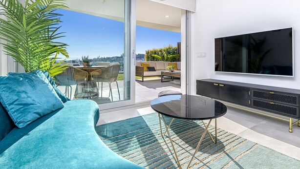 Ein modernes Wohnzimmer mit einem gebogenen, türkisfarbenen Sofa, einer Glastür zum Balkon, einem Flachbildfernseher und einem kleinen runden Tisch.