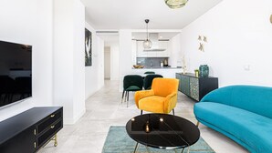 Ein modernes Wohnzimmer mit einem schwarzen Couchtisch, einem türkisfarbenen Sofa und einem gelben Sessel. Im Hintergrund ist eine Küche zu sehen.