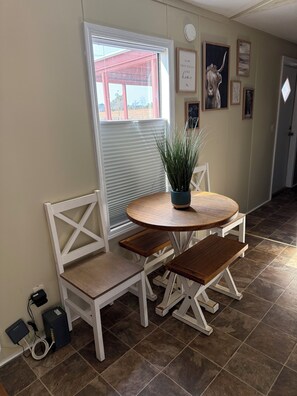 Un coin repas avec une table ronde en bois, deux bancs blancs rembourrés, une plante en pot et une fenêtre avec des stores.
