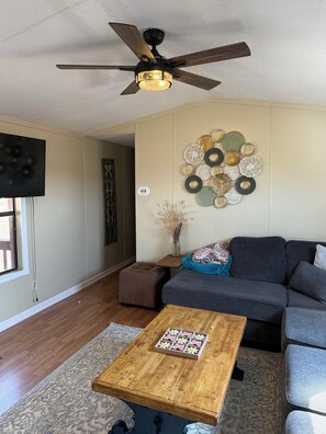 Un salon avec un ventilateur de plafond, un canapé gris, une table basse en bois, un tapis à motifs et un élément décoratif mural.