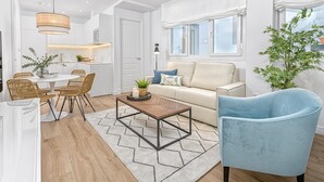 A modern living room with a sofa, a coffee table, and a blue armchair. There is a dining area with wicker chairs and a table. The room has white walls and a wooden floor. - Bright apartment in Málaga (Málaga)
