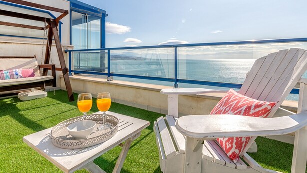 Eine Terrasse mit Tisch, zwei Stühlen und einer Schaukel. Die Terrasse bietet Blick auf ein Gewässer.