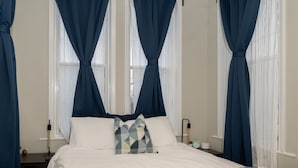 Un lit bien fait, avec une couette blanche et des oreillers, une table de chevet avec une lampe, et une fenêtre ornée de rideaux légers.