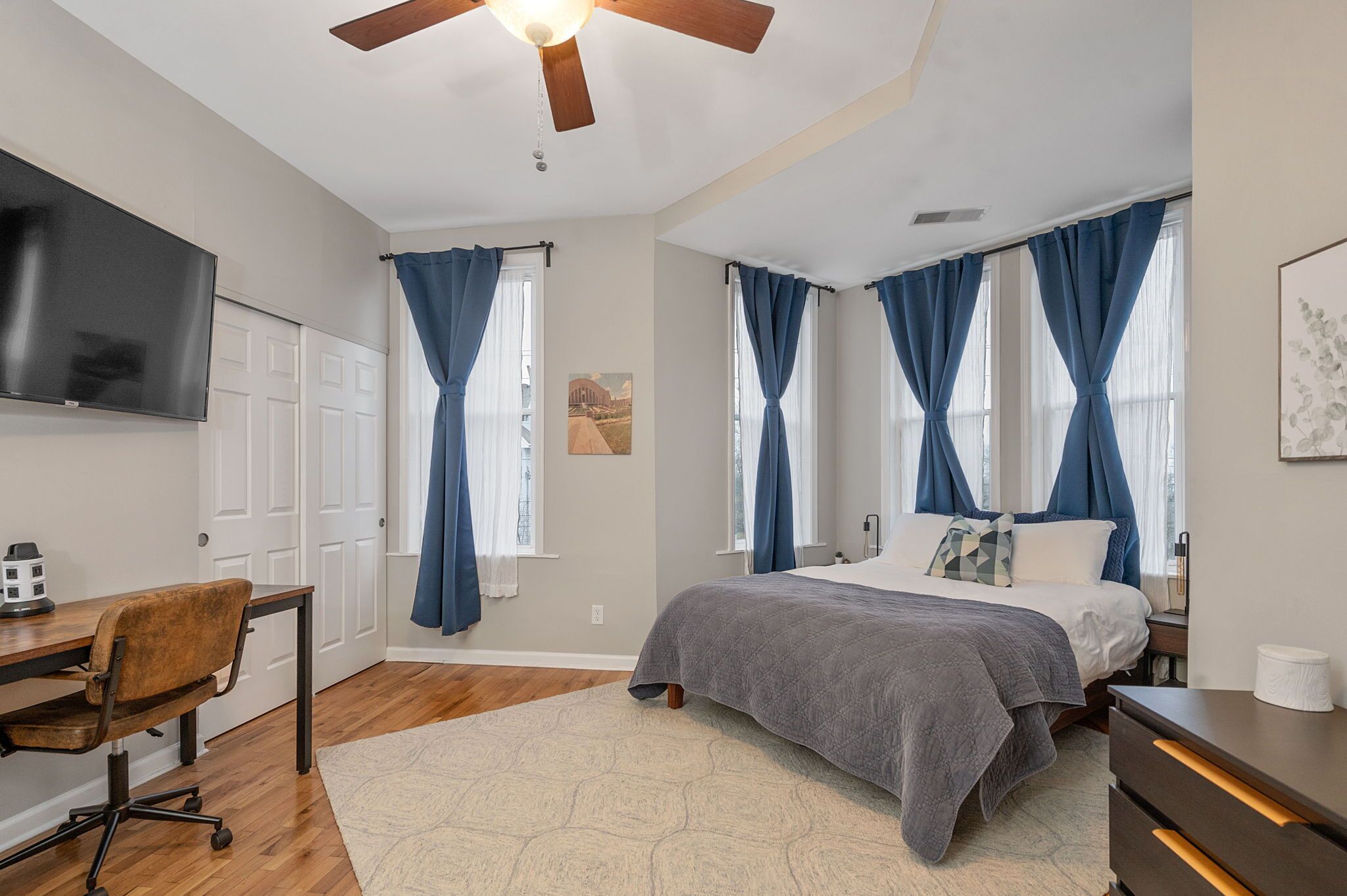 A bedroom with a bed, a desk, a chair, a TV, and a ceiling fan.