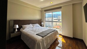 Camera d'albergo con un letto grande, due comodini con lampade, vista sugli edifici attraverso un'ampia finestra e un soffitto con illuminazione a incasso.