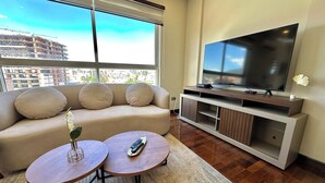 Un soggiorno moderno con una grande televisione a schermo piatto, un divano beige con tre cuscini, due tavolini rotondi e vista sulla città attraverso ampie finestre.