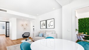 Een moderne woonkamer met een ronde witte tafel, teal stoelen en een grijze bank. Er hangen kunstwerken in lijsten aan de muur en een groene wandtuin.