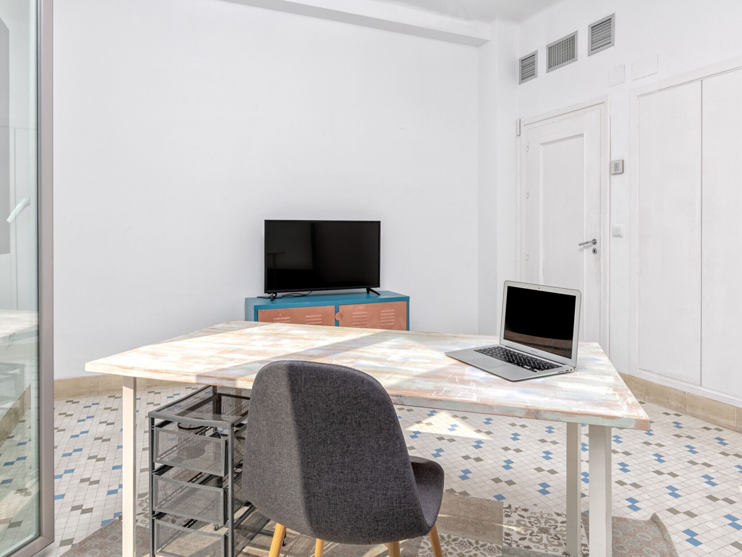 Una habitación pequeña con suelo de baldosas, un escritorio con una computadora portátil, una silla, un televisor sobre un mueble y una puerta.