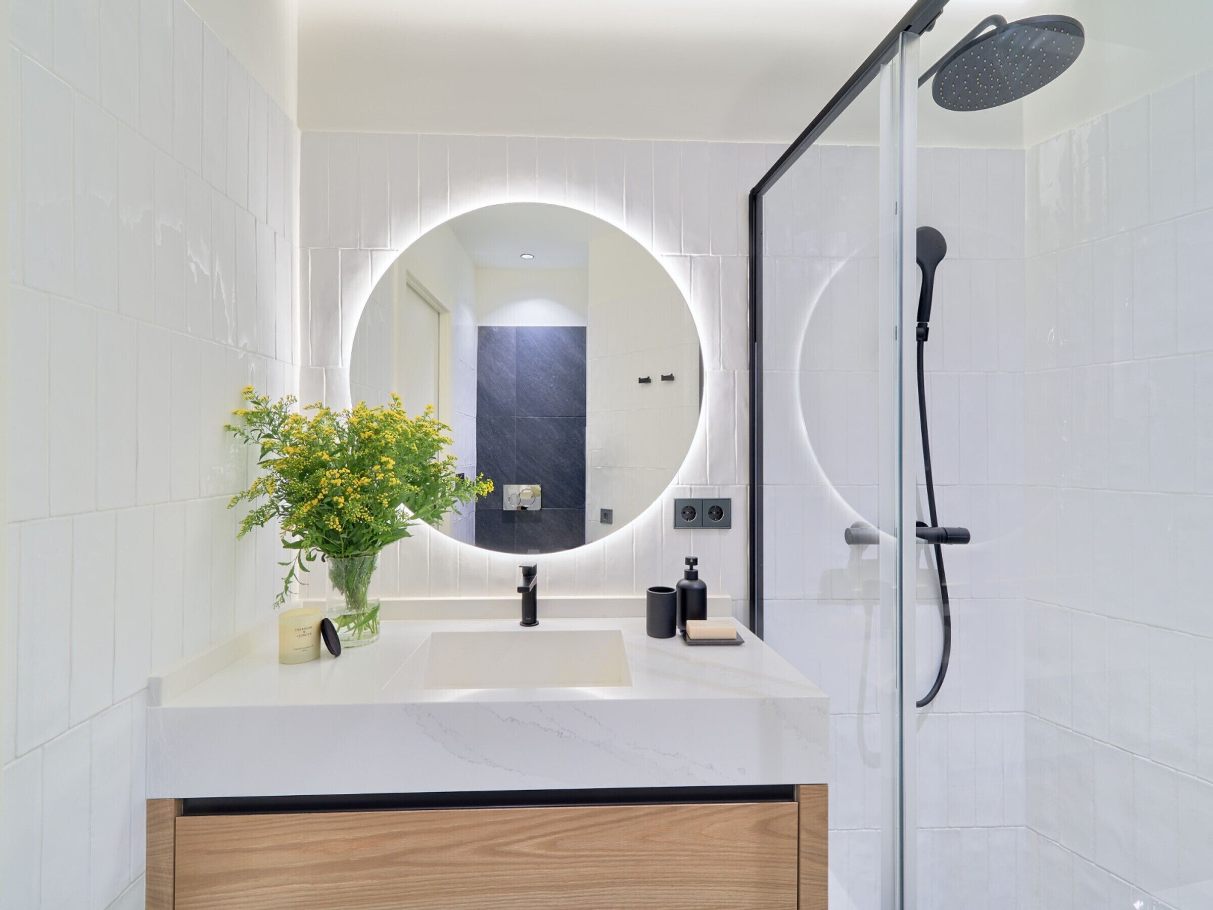 Une salle de bain moderne avec un miroir rond, des carreaux blancs, un meuble en bois, un lavabo, une douche et un vase avec des fleurs jaunes.