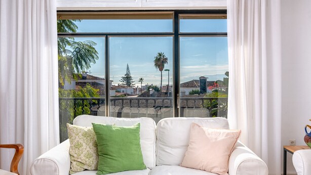 Un sofá de cuero blanco con cojines verdes y rosas, una mesa de centro de madera con objetos decorativos, y un ventanal con vistas a la ciudad.