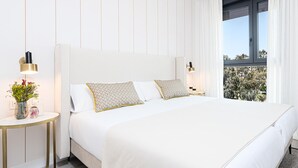 Ein modernes Hotelzimmer mit einem großen Bett, weißer Bettwäsche, einem runden Beistelltisch mit Blumen und Blick auf Bäume durch das Fenster.