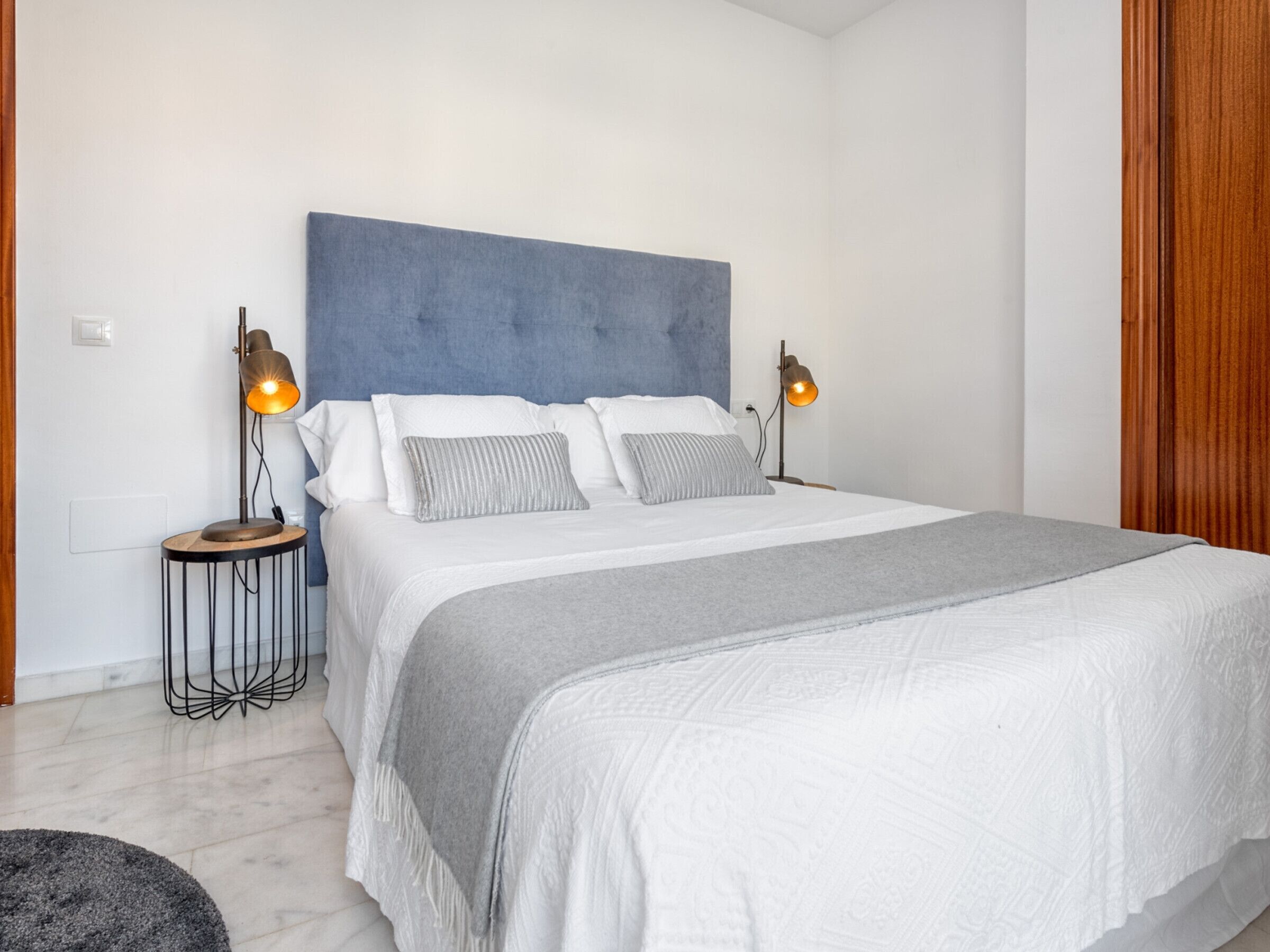 Ein Schlafzimmer mit einem großen Bett, zwei Nachttischlampen, einer grauen Tagesdecke und einem blauen Kopfteil.