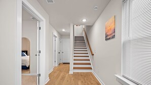 Un pasillo con suelo de madera, paredes blancas, una escalera de madera y una puerta que da a un dormitorio.