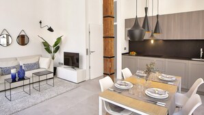 Ein modernes Wohnzimmer mit einer Couch, einem Couchtisch und einem Fernseher. Eine Essecke mit Holztisch und Stühlen ist ebenfalls zu sehen.