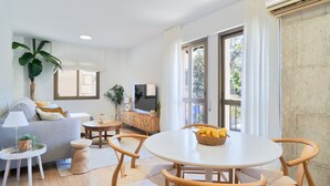 Un soggiorno moderno con un divano grigio, un tavolo da pranzo rotondo con sedie in vimini, una TV su un mobile in legno e un'ampia finestra con tende.