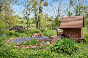 Een rustieke houten chalet met een klein terras, omgeven door een tuin met een vijver en diverse planten.