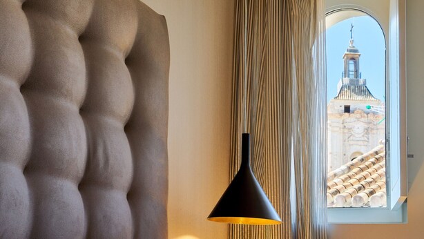 Une chambre dotée d’une tête de lit capitonnée, d’une suspension noire et offrant une vue sur un bâtiment à travers une fenêtre à cadre blanc.